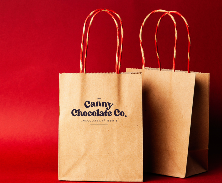 Two brown paper bags with 'Canny Chocolate Co.' branding on a red background